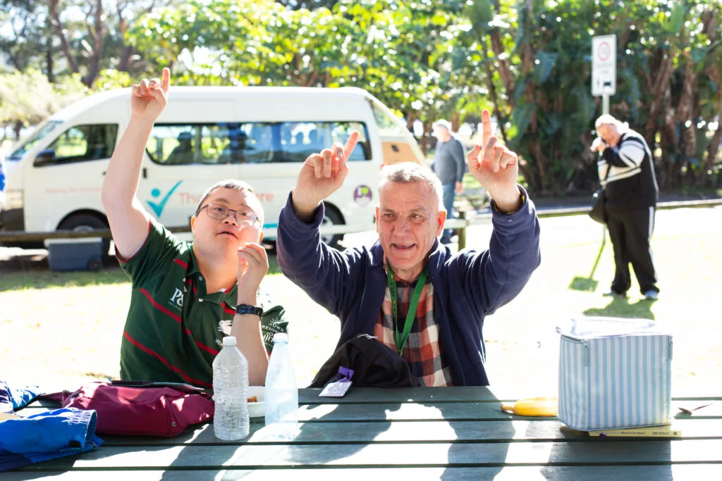 Two Therapy Care day program participants cheering at a picnic table on a beach outing
