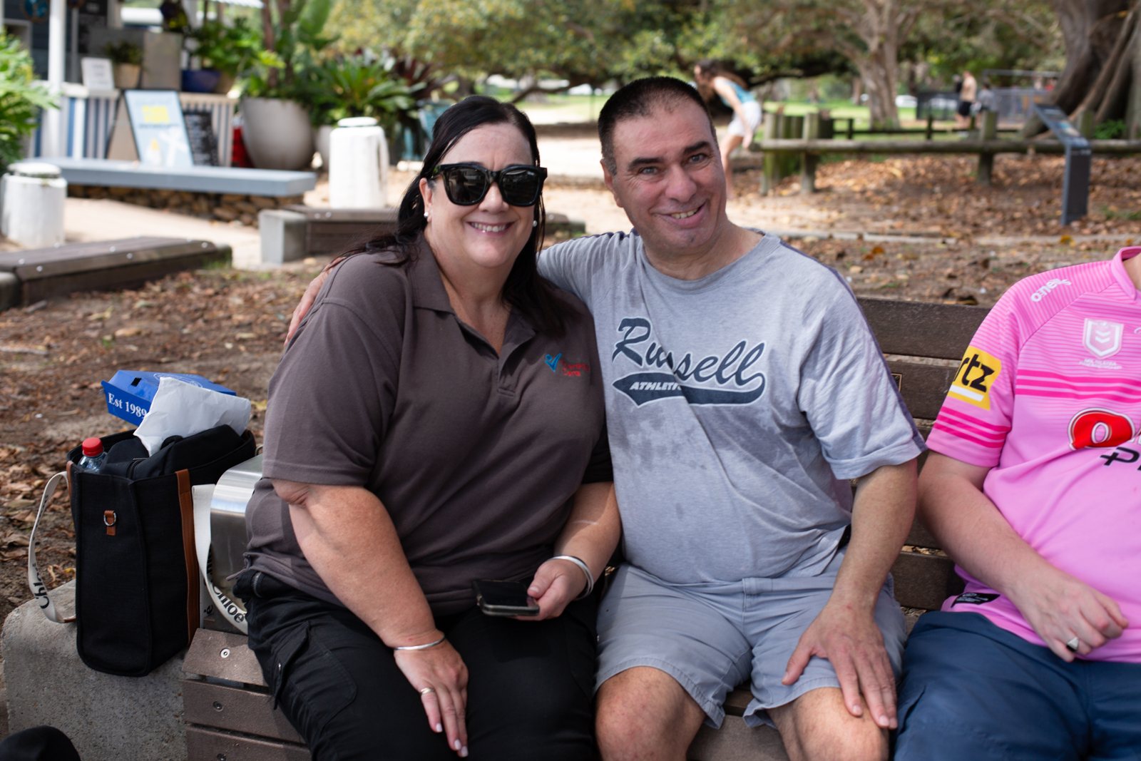 A Therapy Care support worker and a participant sitting together on a park bench, smiling, during a day program outing